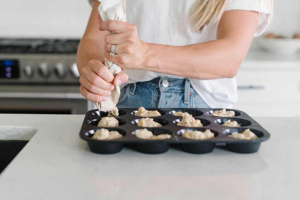 Person filling a muffin tin with batter using a piping bag in a kitchen.