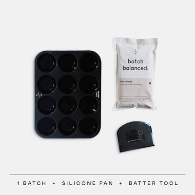 Black silicone muffin pan, white bag labeled 'batch balanced', and black batter tool on a light gray background.